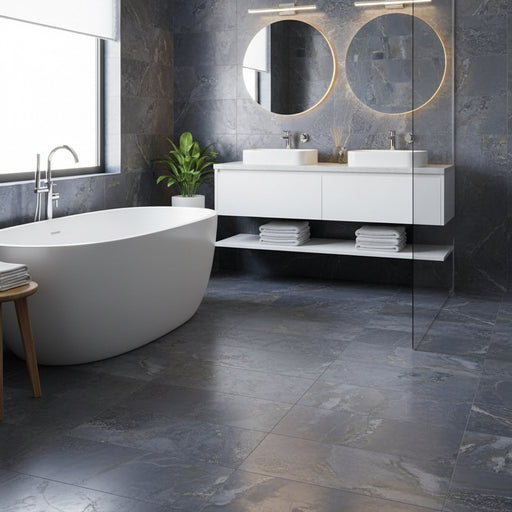 A modern bathroom interior featuring large slate-look tiles in blue, gray, and gold veining on the wall and floor, a light wood platform, a white freestanding bathtub, and a minimalist bench with folded towels.