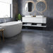 A modern bathroom interior featuring large slate-look tiles in blue, gray, and gold veining on the wall and floor, a light wood platform, a white freestanding bathtub, and a minimalist bench with folded towels.